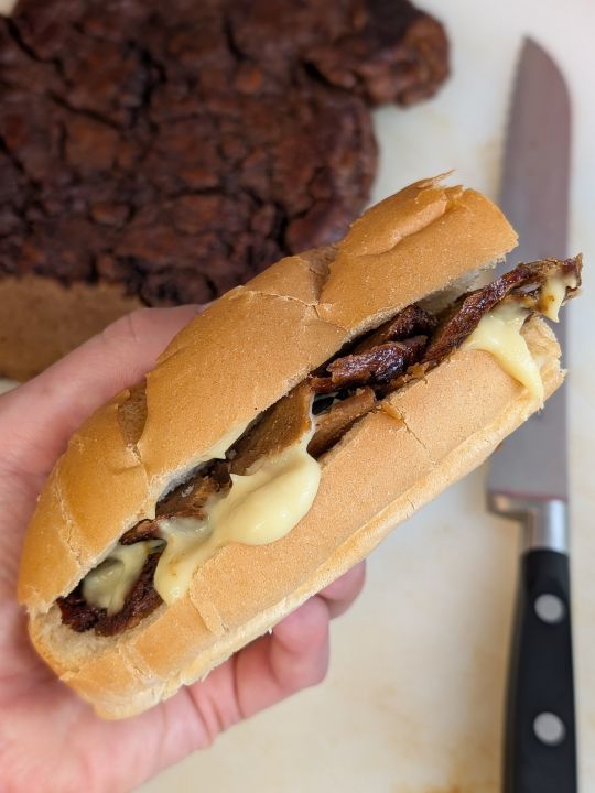 hand holding a vegan cheesesteak above a cutting board with a slab of flank-style vegan steak next to a serrated knife